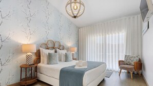 Un dormitorio con una cama, dos mesitas de noche, una silla, un televisor y una ventana con cortinas.