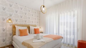 Een net opgemaakt bed met kussens, een bedlampje en een raam met dunne gordijnen.