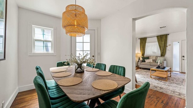 A dining area with a wooden table, green upholstered chairs, a woven pendant light, and a vase with flowers.