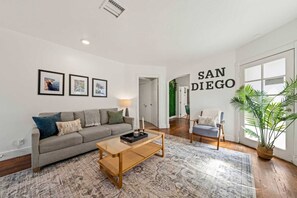 Una sala de estar moderna con un sofá gris, una mesa de centro de madera y una silla. Hay cuadros enmarcados en la pared, una lámpara y un ventanal con una planta.