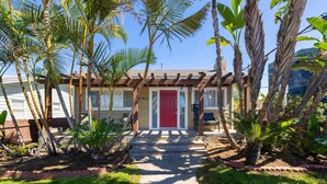 A house with a red door, a covered porch, and a well-maintained garden with palm trees.