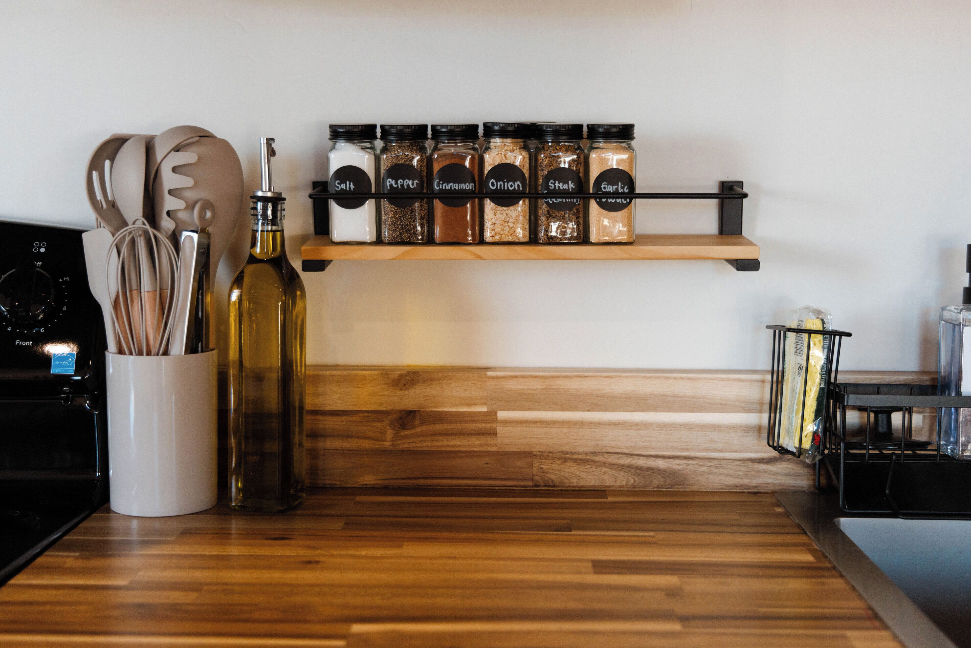 A kitchen counter with a wooden floor, a spice rack with labeled jars, a white container with utensils, and a bottle of oil.