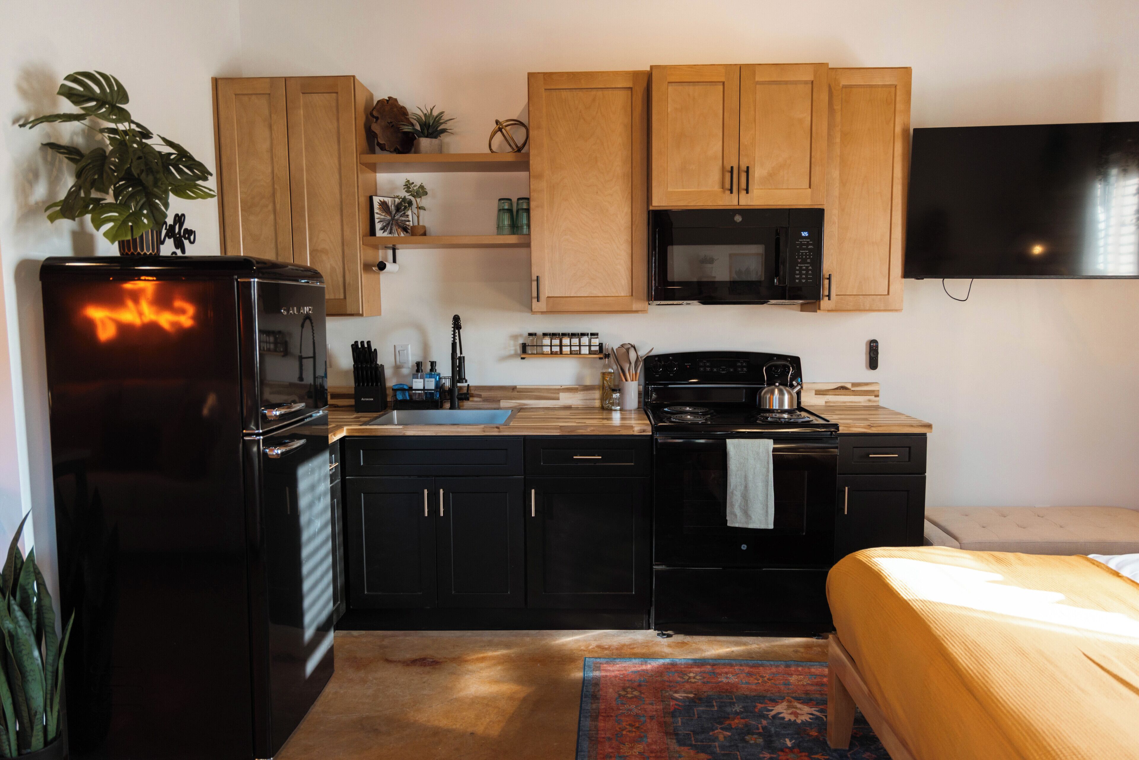 Una cocina compacta con electrodomésticos negros, armarios de madera, un microondas y un televisor de pantalla plana.