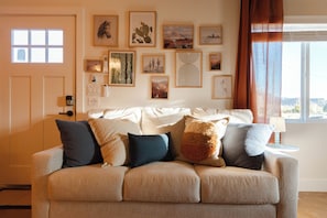 Un salon avec un canapé beige, divers coussins, un mur orné de tableaux encadrés, une porte et une fenêtre avec des rideaux.