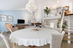 Een eetruimte met een ronde tafel, rieten stoelen, een kroonluchter en een trap naar een hoger niveau.