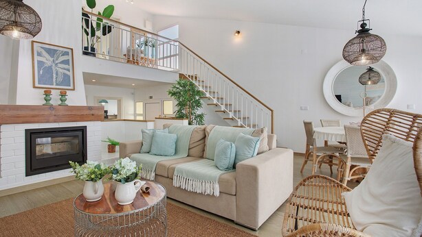 Un salon avec une cheminée, un canapé, une table basse, des fauteuils en osier et un escalier.