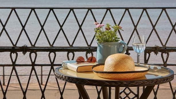 Een tafel met een boek, een hoed, een glas water en een potplant op een balkon met een metalen hekwerk en uitzicht op de zee.