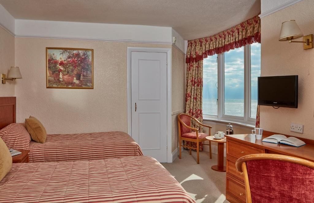 Habitación de hotel con dos camas, un escritorio con silla, televisión, ventana con vistas y un cuadro en la pared.