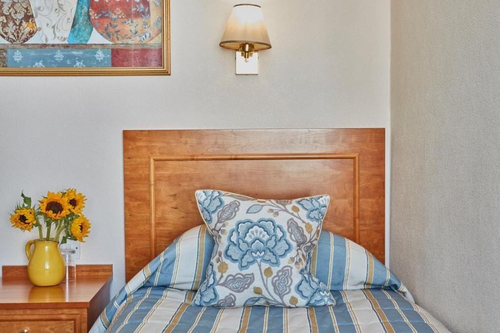 Una cama bien hecha con cabecera de madera, un cojín decorativo, una mesita de noche con un jarrón de girasoles y una lámpara de pared.