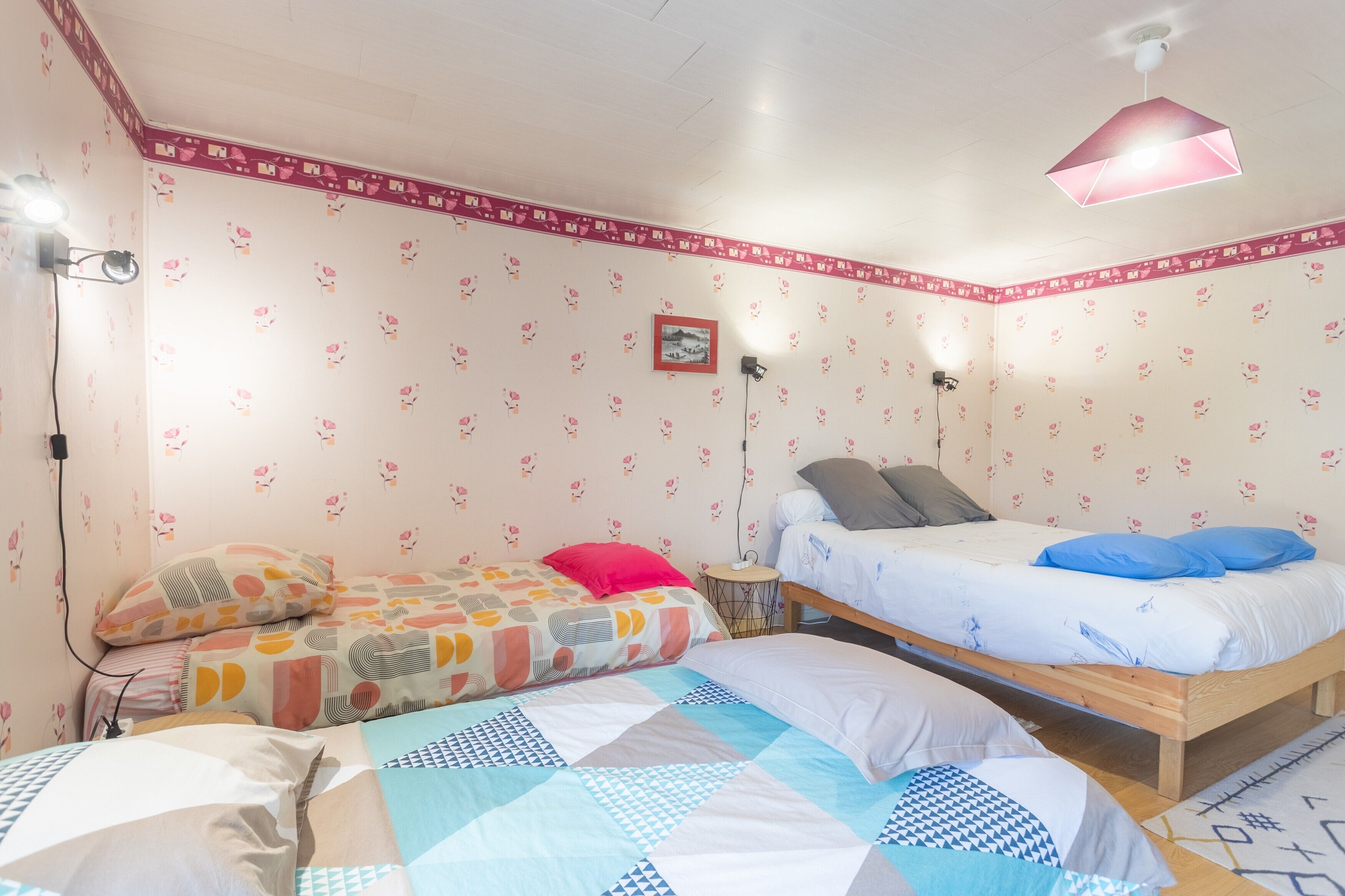 A bedroom with two beds, a small bedside table, a wall-mounted lamp, and a ceiling light fixture.