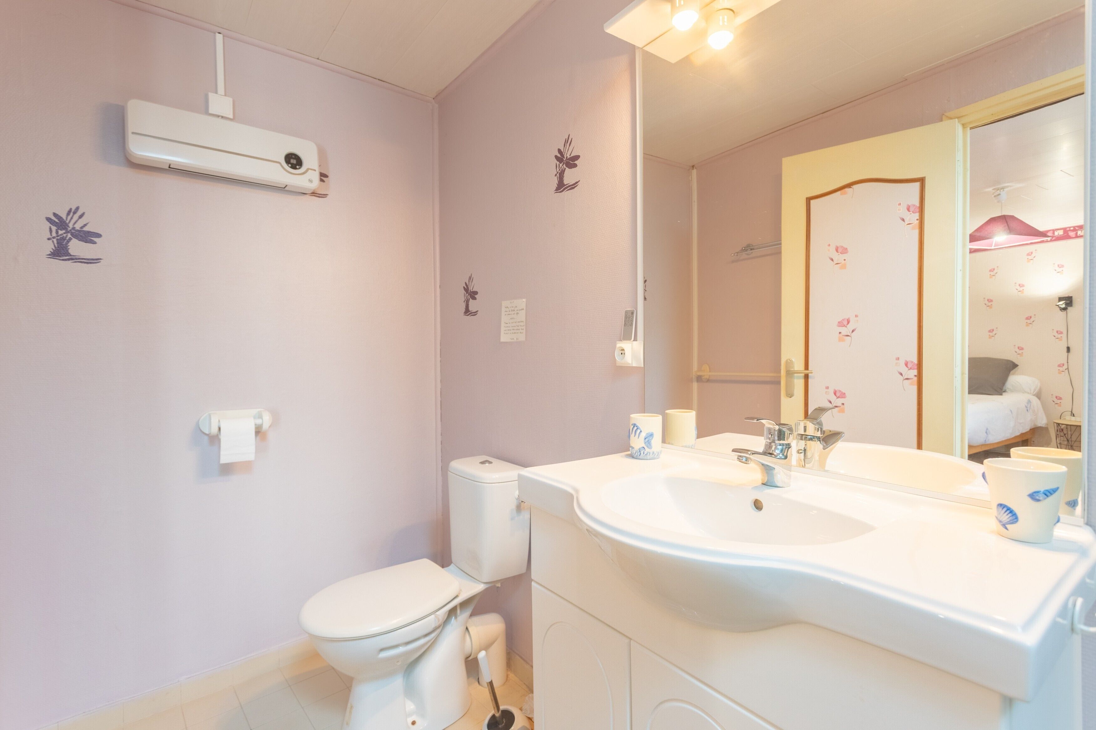 A bathroom with a toilet, sink, mirror, and a wall-mounted air conditioner.