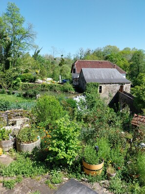 Une maison de campagne en pierre, avec un toit rouge, entourée d’une végétation luxuriante et d’un jardin où poussent diverses plantes et fleurs.