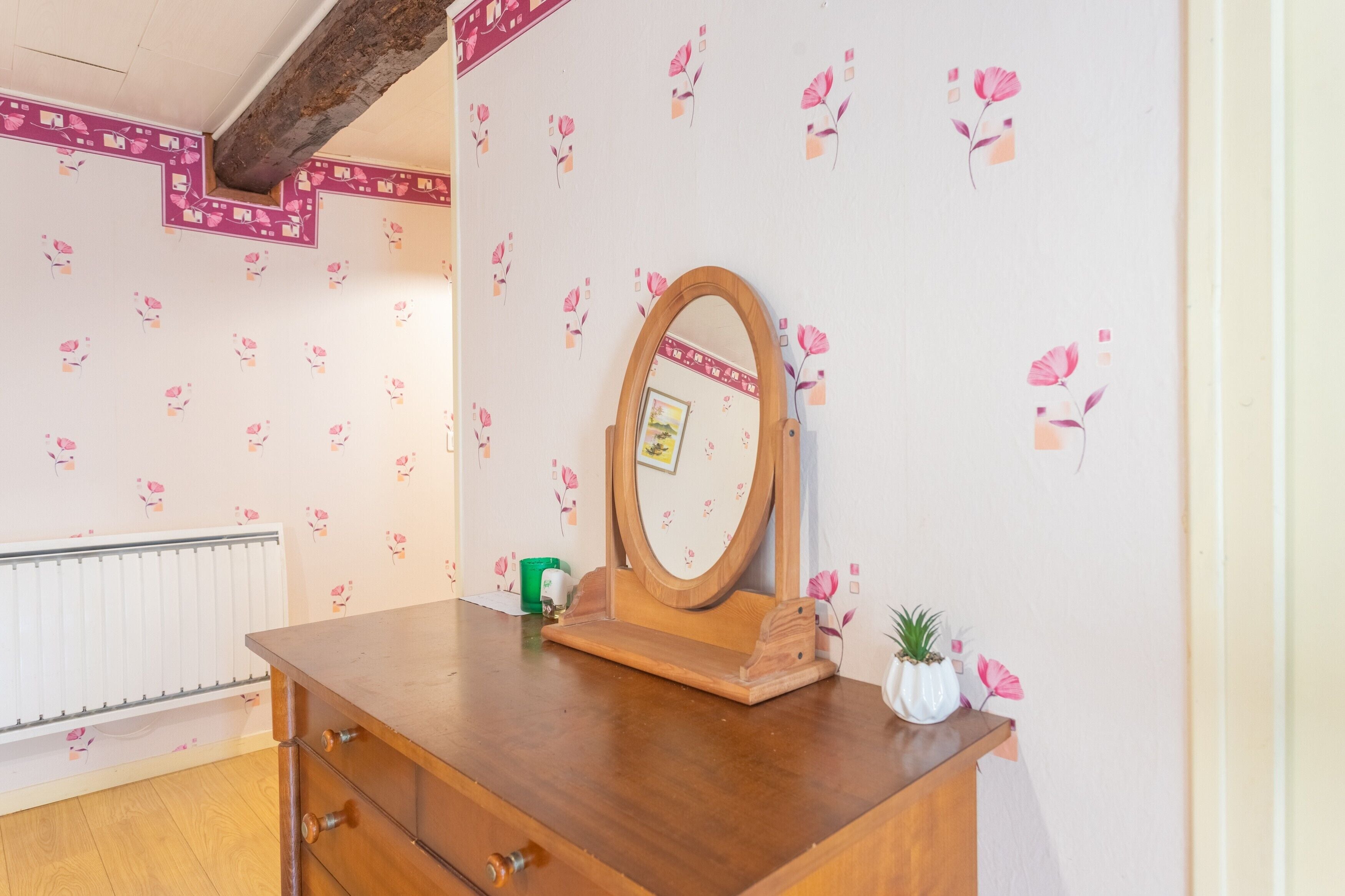 Une commode en bois avec un miroir, une petite plante et un radiateur, dans une pièce dont le papier peint est à motifs floraux.