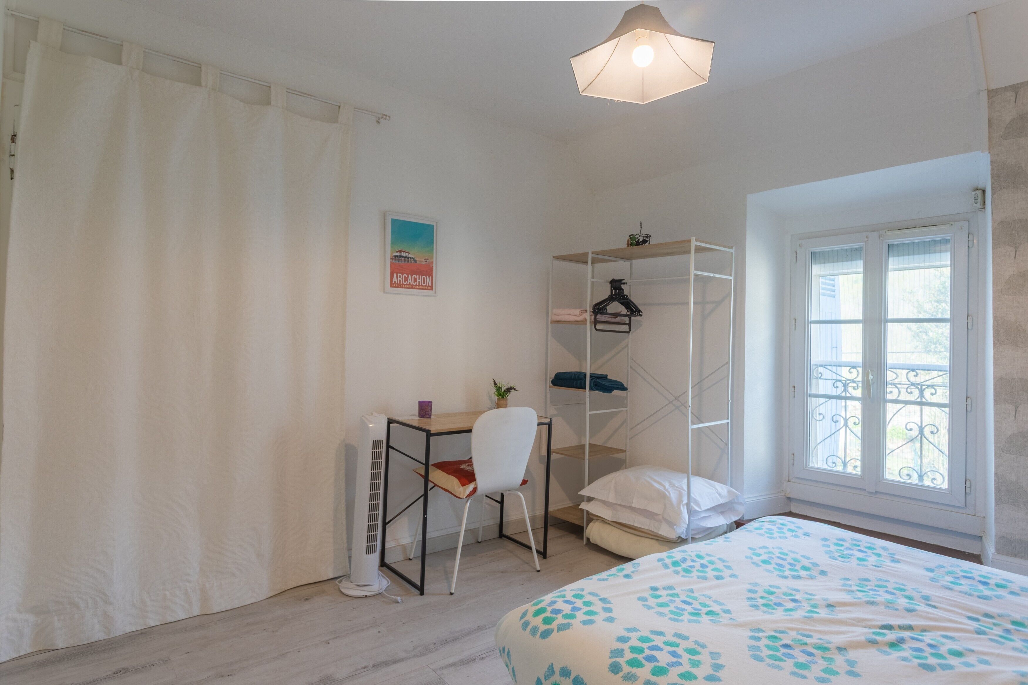 A bedroom with a bed, a desk, a chair, a radiator, a shelf, a window, and a curtain.
