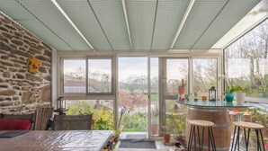 Une pièce avec un mur en pierre, un lit, une table recouverte d’une natte, un tabouret en fût de bois et une vue sur un jardin, visible à travers de grandes fenêtres.