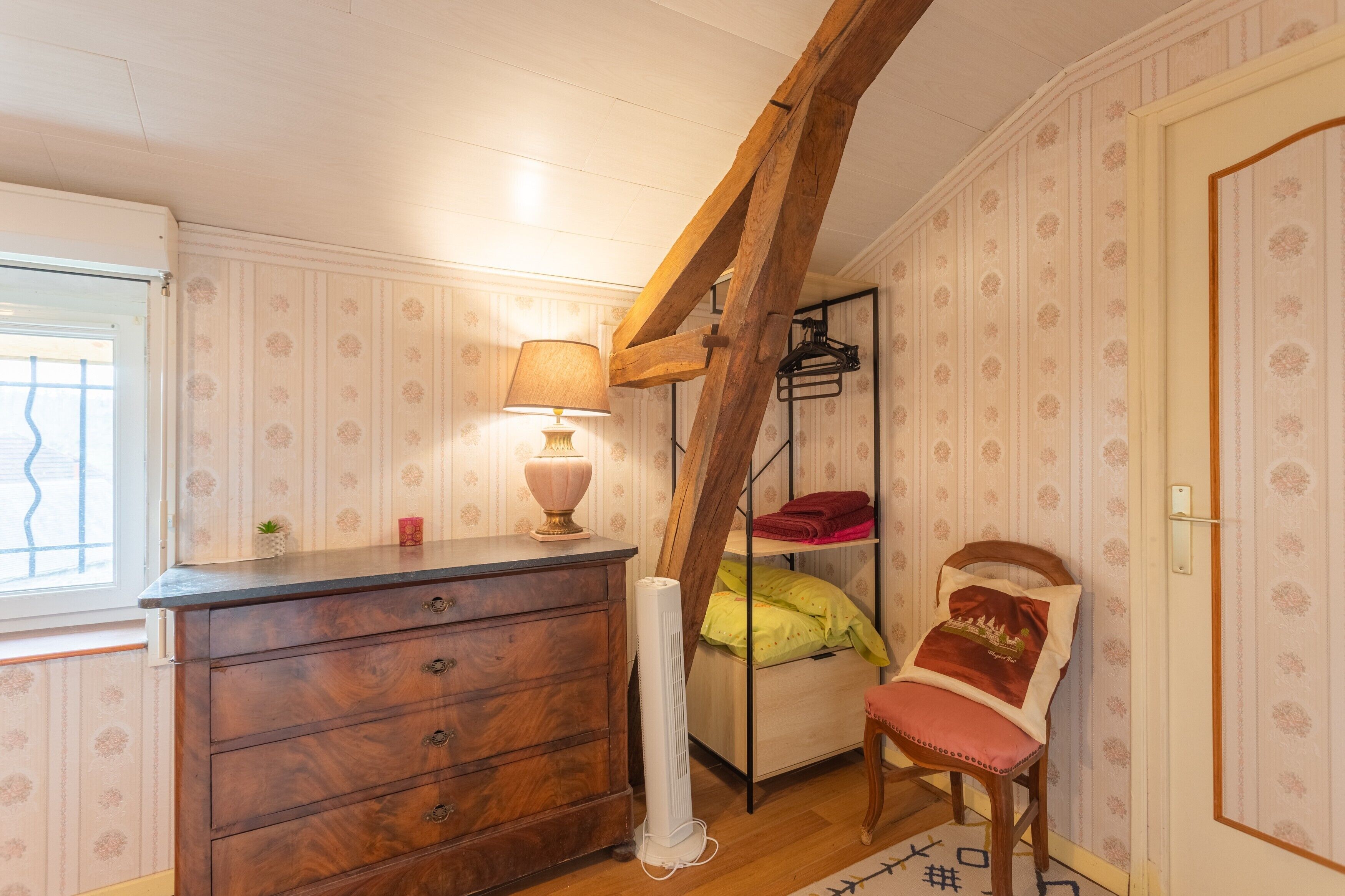 Una habitación con un aparador de madera, una cama con almohadas, una silla, una lámpara y una ventana.