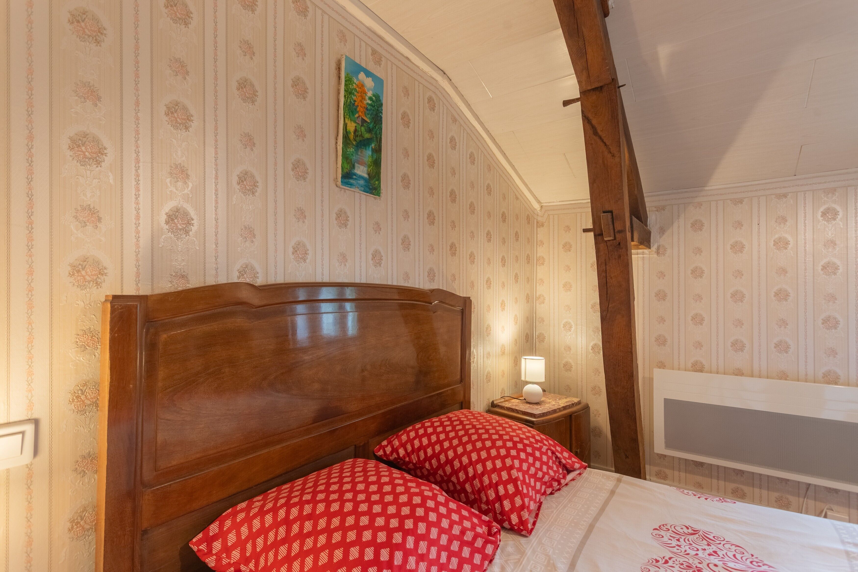Een slaapkamer met een houten hoofdbord, een bed met rode, gemêleerde kussens, een nachtkastje met een lamp en een schilderij aan de muur.
