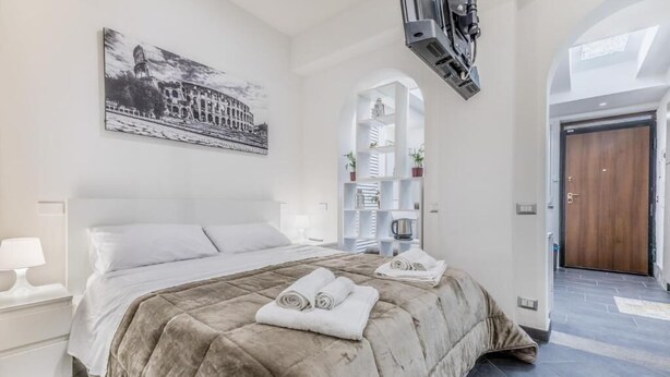 A hotel room with a large bed, a painting of a historical building, a built-in shelf with plants, and a door leading to another room. - Battistini Sweet House (Roma)