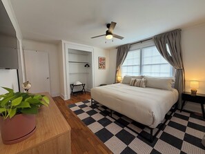 Ein Hotelzimmer mit einem Bett, einem Deckenventilator, einem Fenster mit Vorhängen, einer Nachttischlampe, einem karierten Teppich und einer Topfpflanze.