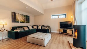 A living room with a sectional sofa, a flat-screen TV, a fireplace, and a large window. - The Stables (Huntingdon)
