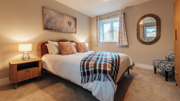 A bedroom with a bed, bedside table, lamp, window with curtains, mirror, and a chair. - The Stables (Huntingdon)