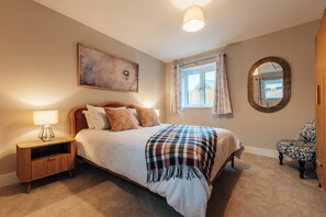 Ein Schlafzimmer mit einem Bett, Nachttisch, Lampe, Fenster mit Vorhängen, Spiegel und einem Stuhl.