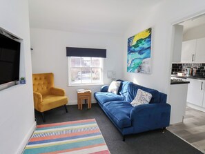 A compact living space with a sofa, a yellow armchair, a small table, a colorful rug, a painting, and a kitchen area. - Granby Place Apartment 3 (Scarborough)