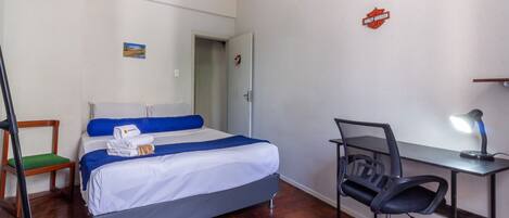 A hotel room with a bed, a desk with a chair, a lamp, a door, and a small picture on the wall.