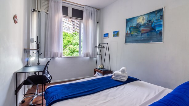 Una camera da letto con un letto, una scrivania, una sedia, una finestra con tende, un quadro e un piccolo tavolo con un asciugamano.