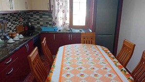 Una cocina con mantel a cuadros, sillas de madera, un refrigerador y un ventanal con cortinas.