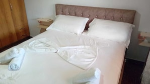 Una cama bien tendida con ropa y toallas blancas, un armario de madera y una mesita de noche con lámpara.