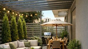 Een terras met een bank, eettafel en stoelen, omgeven door planten en een pergola met lichtslingers.