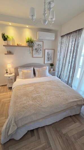 A bedroom with a large bed, a bedside table with a lamp, a wall-mounted air conditioner, and a window with curtains. - Sagma Maisonette with pool and garden  (Glyfada)