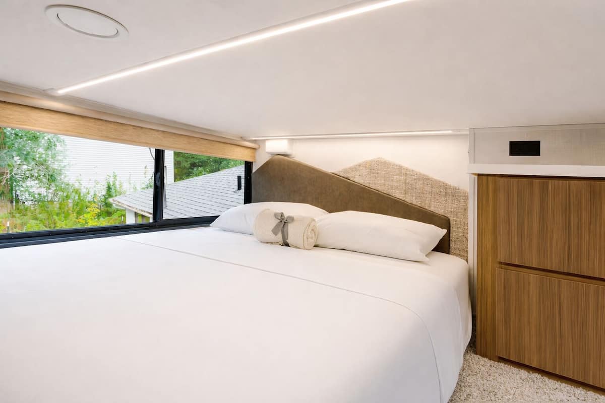 A modern bedroom with a large bed, a wooden headboard, a built-in wardrobe, and a view of greenery outside.