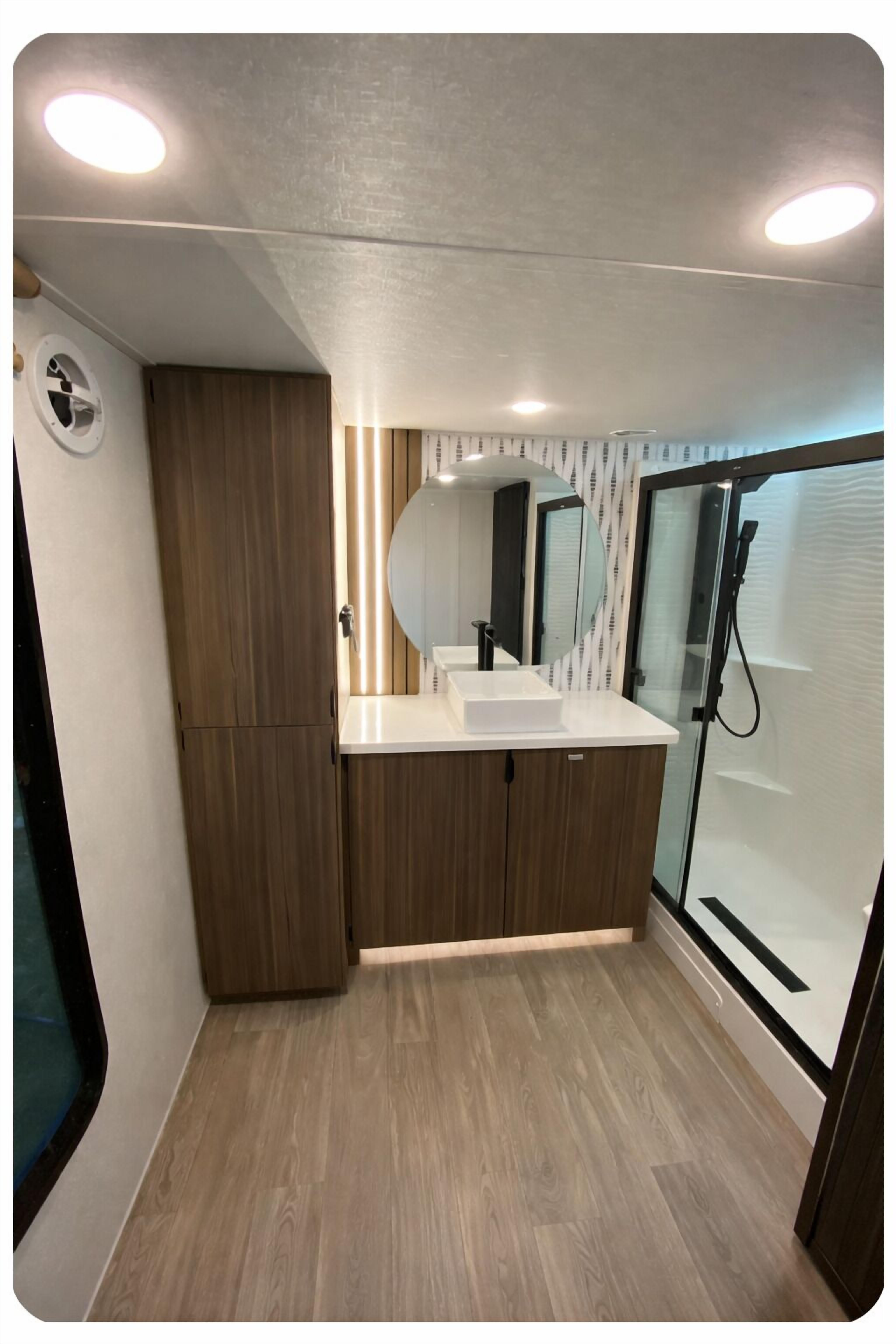A modern bathroom with a wooden vanity, a large mirror, and a glass shower enclosure.