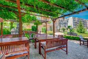Un patio al aire libre con mesas y bancos de madera, rodeado de follaje y vistas a edificios de apartamentos.