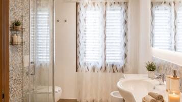 A bathroom with a glass shower enclosure, a toilet, a sink with a mirror, and a window with a white curtain.