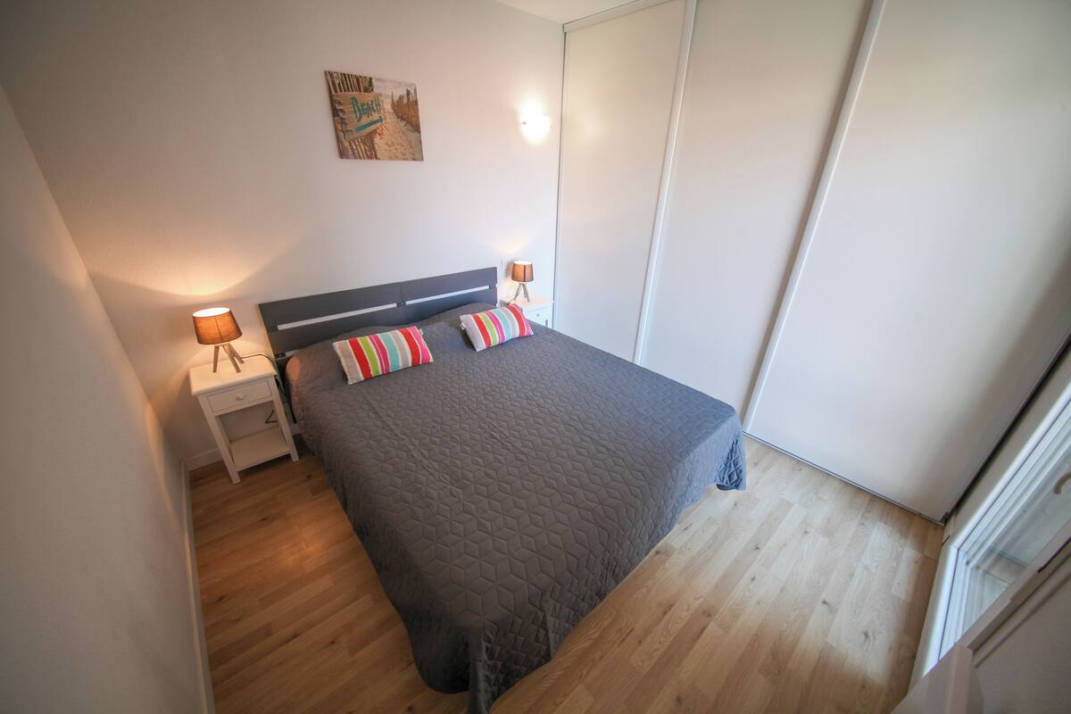 A bedroom with a bed, two bedside tables, a nightstand, a wall-mounted lamp, and a door.