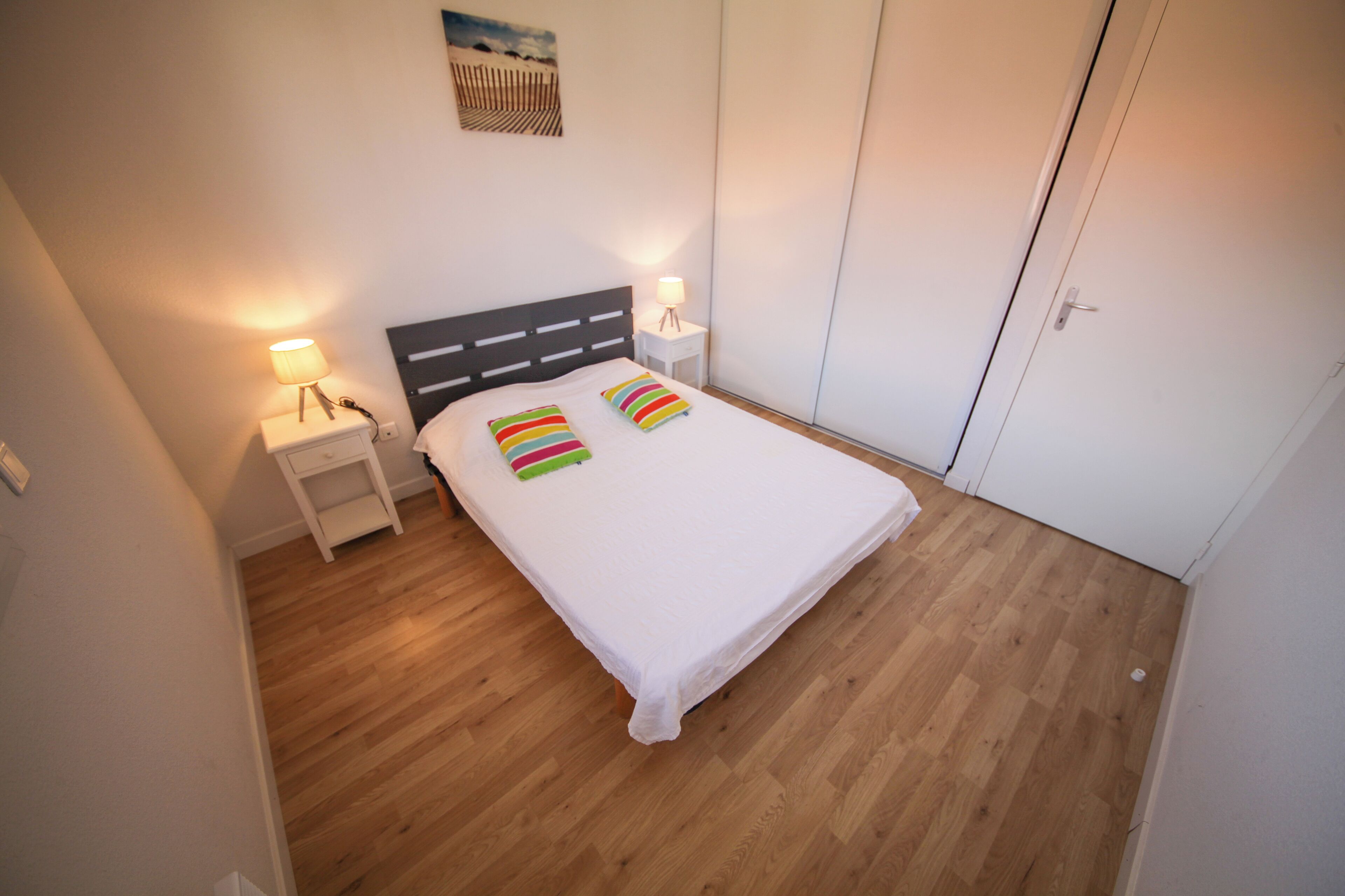 Un lit simple avec un matelas blanc et deux oreillers à rayures, une table de chevet avec une lampe et une armoire dans une pièce dont le sol est recouvert de parquet.