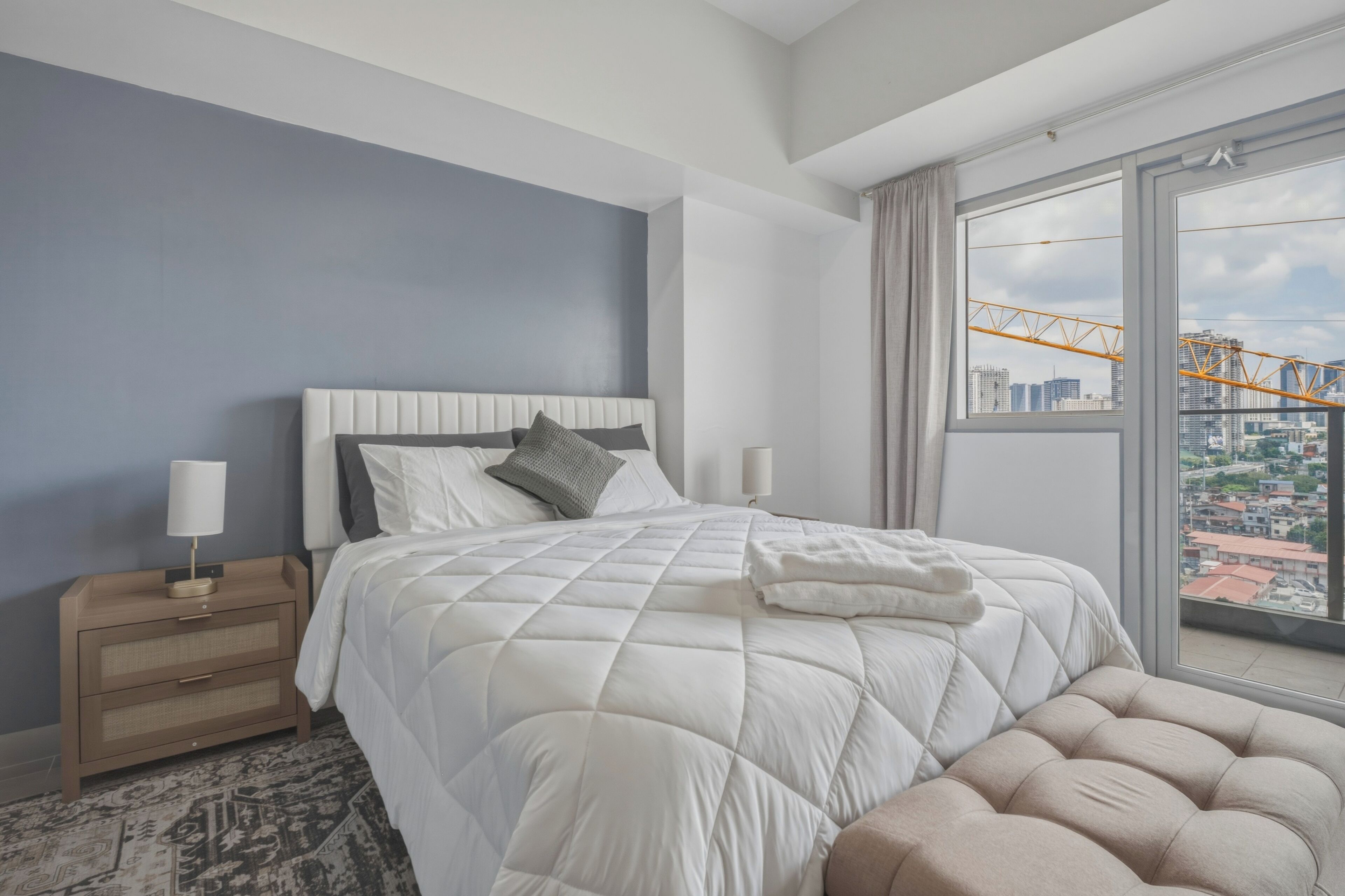 A modern hotel room with a large bed, a nightstand with a lamp, a view of a cityscape through a large window, and a balcony with a railing.