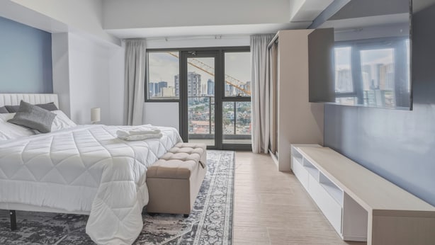 Ein modernes Schlafzimmer mit einem großen Bett, einem Fernseher, einer Bank und Blick auf die Stadt.