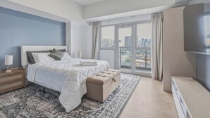 Ein modernes Schlafzimmer mit einem großen Bett, einem Nachttisch, einer Bank, einem Fernseher und Blick auf die Stadt.