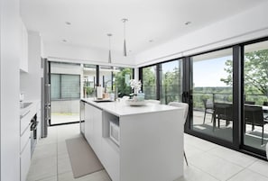 A modern kitchen with a central island, large sliding glass doors leading to a balcony, and a view of greenery outside. - Luxury 2BR Waterfront Condo w/ Garage Parking (Laval)