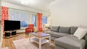 Ein Wohnzimmer mit Fernseher, einer Couch, einem roten Sessel, einem Essbereich mit Tisch und Stühlen sowie großen Fenstern mit Vorhängen.