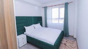 Ein Schlafzimmer mit einem grünen, gepolsterten Kopfteil, weißer Bettwäsche, einem kleinen Nachttisch und einem Fenster mit Vorhängen.