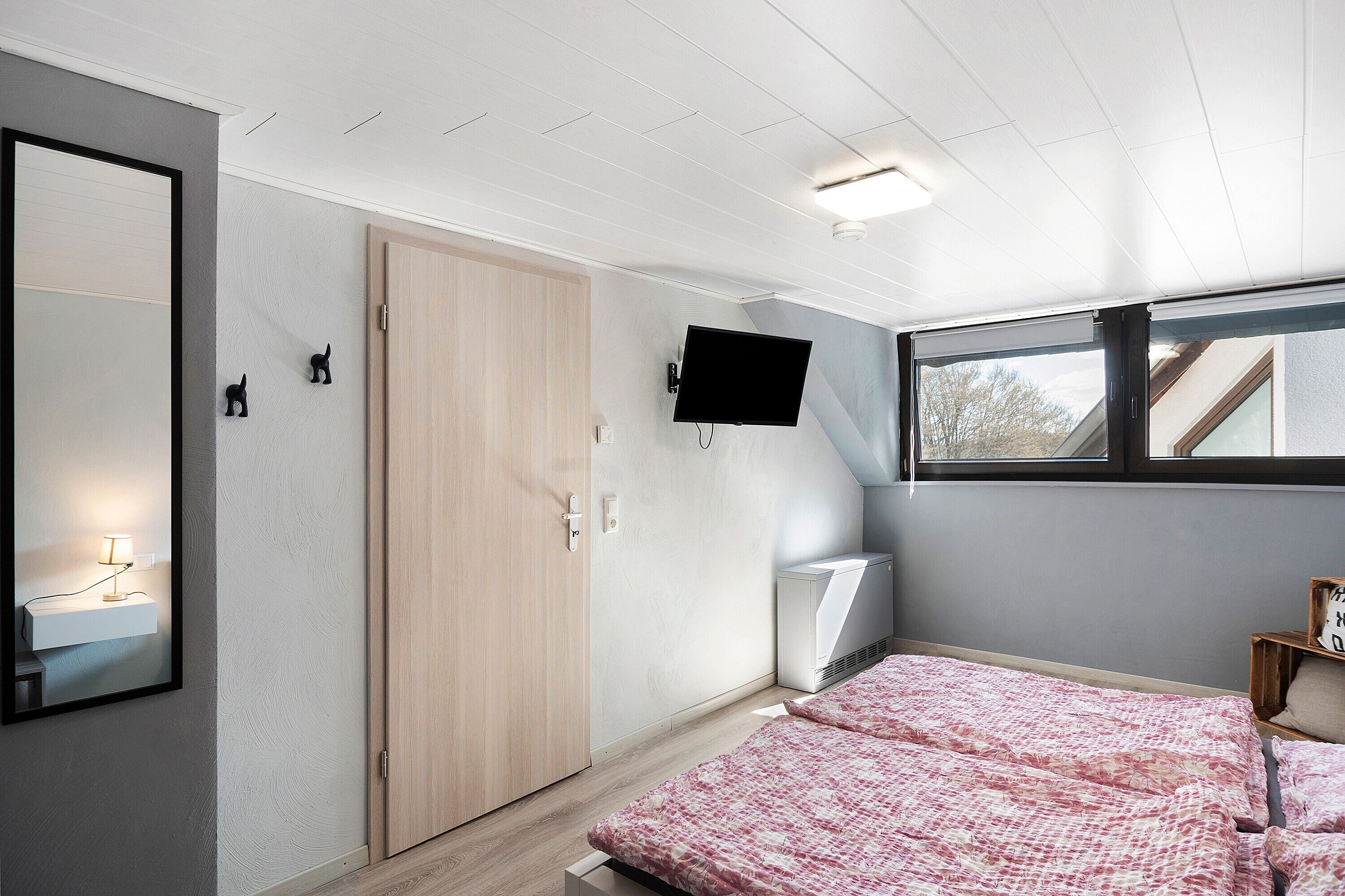 A bedroom with a bed, a TV mounted on the wall, a wardrobe, and a window.