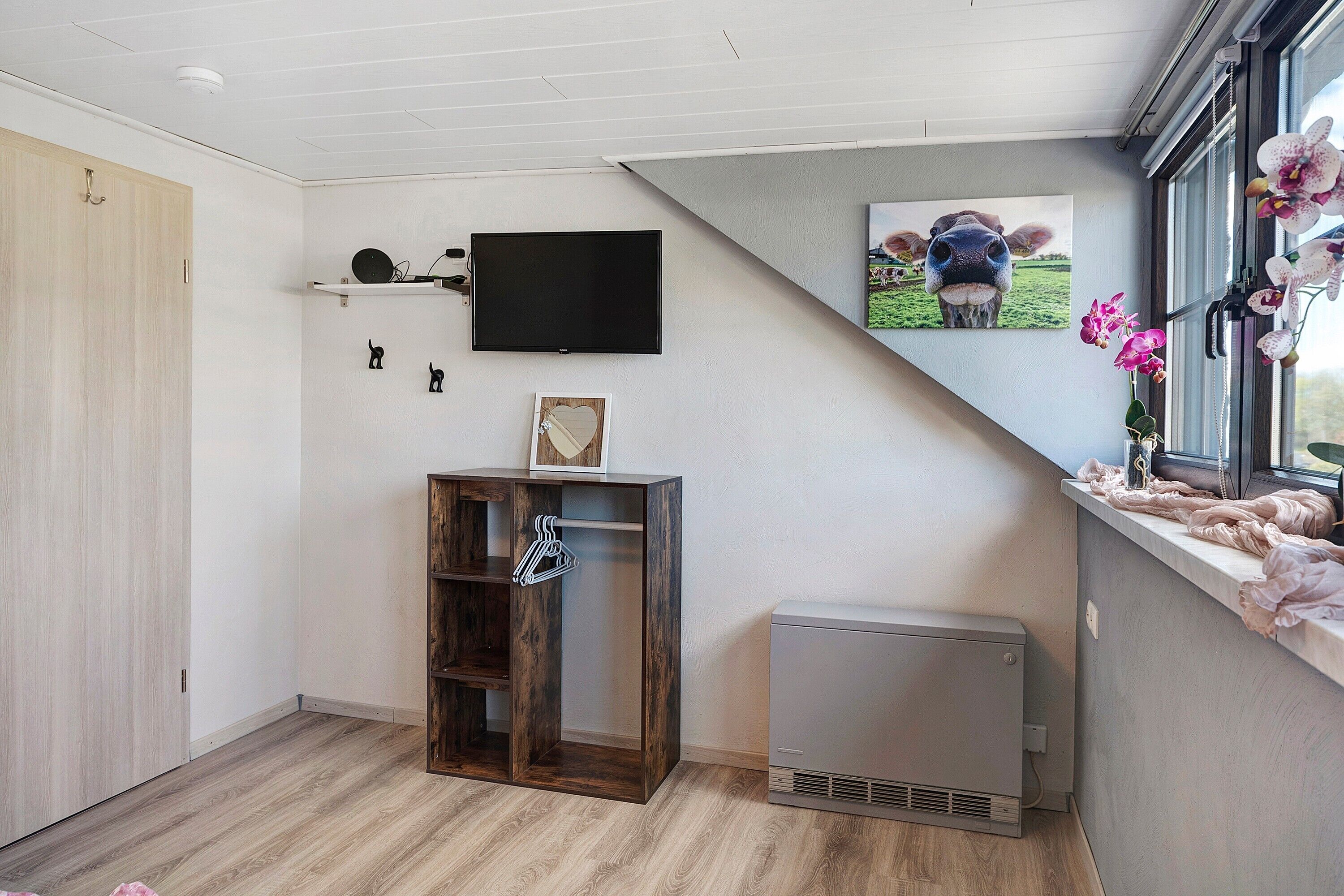 Een kamer met een aan de muur bevestigde televisie, een houten kast, een verwarming en een raam met uitzicht op het landschap.