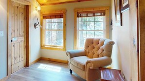 Una habitación con piso de madera, un sillón capitonado, dos ventanas con persianas y una puerta de madera.