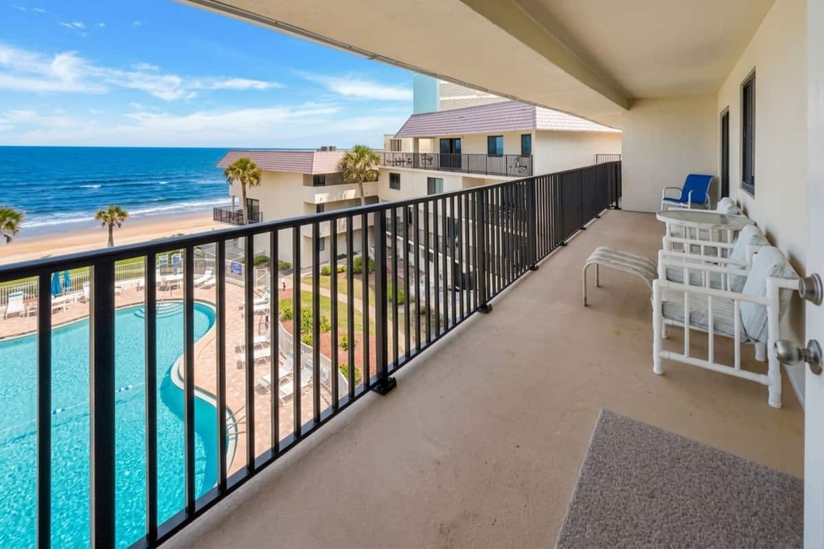 A balcony with a view of a swimming pool and beach, a white chair, and a small table.
