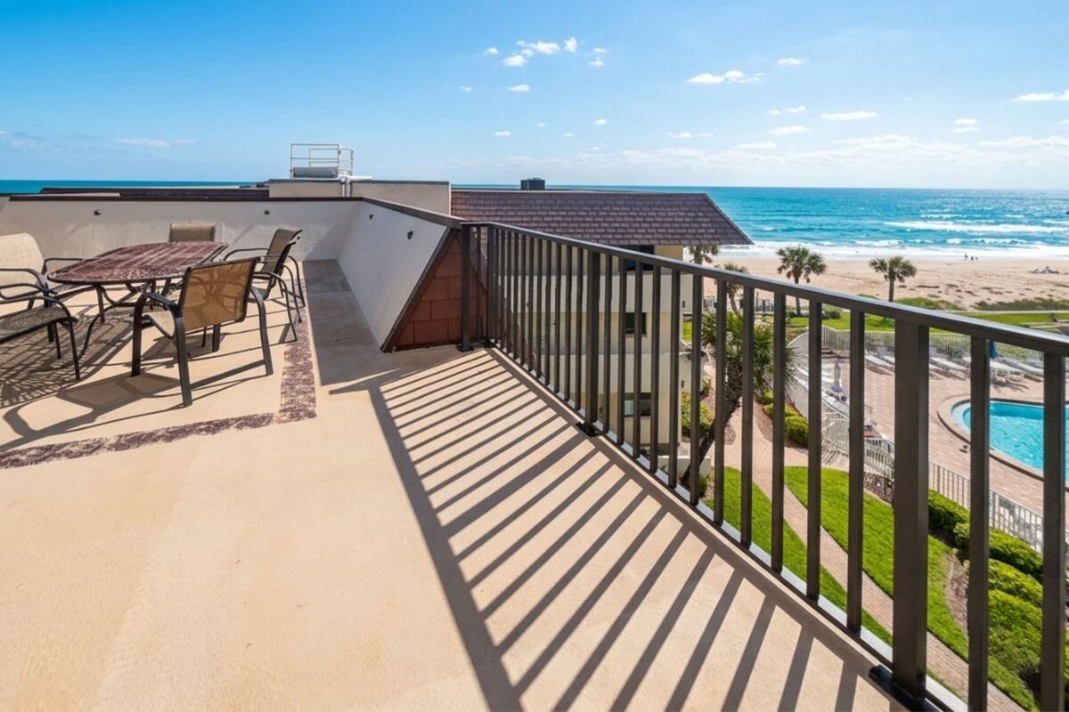 A balcony with a table and chairs, a pool, and a view of the beach and ocean.